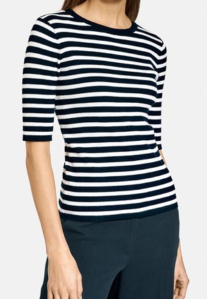 Femme portant un haut côtelé à manches courtes rayé horizontalement noir et blanc, associé à un pantalon de couleur foncée, visible des épaules jusqu'aux hanches.