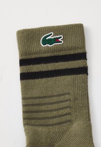 Calcetín verde oliva con dos rayas negras y un logo de cocodrilo verde cerca del puño, textura de punto visible sobre un fondo blanco.