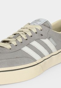Γκρι αθλητικό παπούτσι Adidas Samba με λευκές ρίγες, μπεζ κορδόνια και κρεμ σόλα, σε πλευρική και εμπρόσθια γωνία.