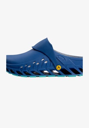 Zapato azul tipo slip-on con parte superior suave, forro de malla y suela texturizada con recortes y un acento en verde azulado. Incluye detalle de logotipo amarillo.