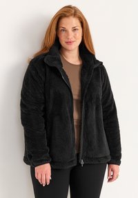 Zwarte fleecjas met een ritssluiting aan de voorkant, hoge kraag en gestructureerde stof. Gedragen over een bruine top en gecombineerd met zwarte broek.
