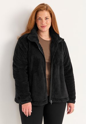 Schwarze Fleecejacke mit einem Reißverschluss vorne, hohem Kragen und strukturiertem Stoff. Über einem braunen Oberteil getragen und mit schwarzen Hosen kombiniert.
