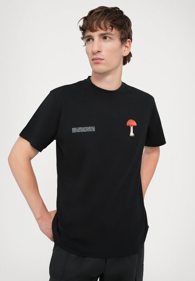 T-shirt en coton noir avec un graphique de champignon rouge et blanc sur la poitrine et du texte blanc sur le côté. Col rond et manches courtes.