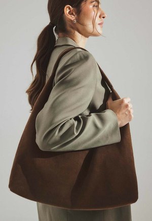 Rjava semišasta torbica za ramo z mehko strukturo in širokim odprtjem, ki jo nosi oseba v svetlo zeleni blazerji, nagnjena na stran.