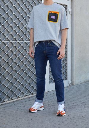 Camiseta gris de gran tamaño con un bolsillo de malla negra y cremallera amarilla, combinada con jeans azul oscuro y zapatillas blancas-naranjas. Fondo texturizado.