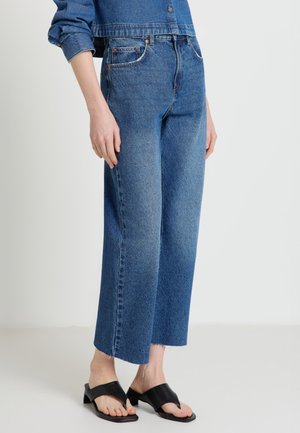 Personne portant un jean bleu large coupé court avec des ourlets bruts et des sandales à talons noires ouvertes, debout devant un fond uni.