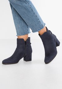 Botines de ante azul marino con tacón cuadrado, cremallera lateral y una textura suave. Presentan un diseño minimalista y una silueta elegante.