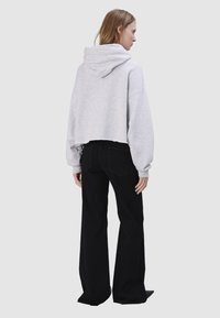 Kobieta w jasnoszarej krótkiej bluzie z kapturem i czarnych szerokich spodniach, stojąca bokiem na neutralnym szarym tle.