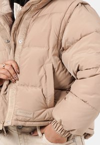 Beige Steppjacke mit gestepptem Muster, auffälligen Nähten und einem Reißverschluss vorne. Verfügt über einen gerippten Bund und eine Markenkennzeichnung am Saum.