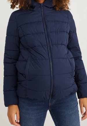 Person iført en marineblå quiltet dunjakke med forreste lynlås og lommer, kombineret med blå denimjeans.