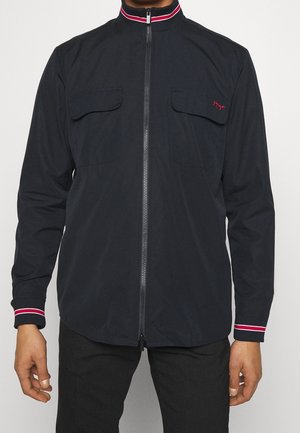 Personne portant une veste noire zippée avec des poignets et un col rayés rouge et blanc, et des poches à rabat sur la poitrine.