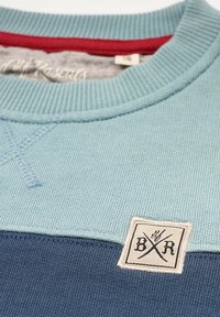 Vaaleansininen neule, jossa on ribbineulostekstuuri, tummansininen alareuna, punainen korostus kaula-aukossa ja merkitty kangaspalikka.