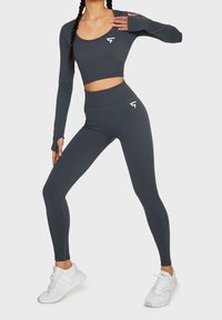Tummanharmaa urheiluasu, joka koostuu pitkähihaisesta crop-topista ja korkeavyötäröisistä legginsseistä. Asussa on logo-yksityiskohtia ja sileä, joustava materiaali.