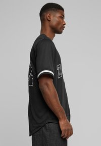 Maillot noir à manches courtes avec des accents à rayures blanches et un lettrage imprimé au dos, offrant une coupe décontractée et une texture lisse.