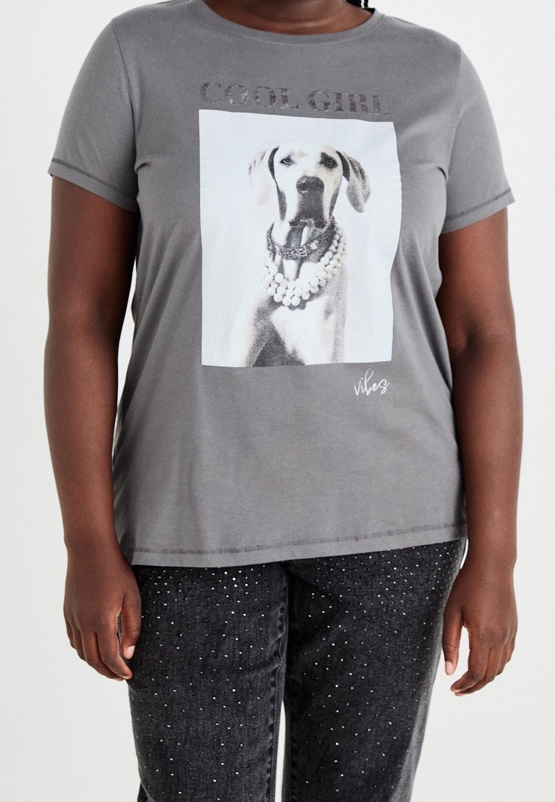 Harmaa t-paita, jossa on koirakuva ja "COOL GIRL" -teksti kiiltävän hopeisena. Lyhythihaisessa paidassa on pyöreä kaula-aukko, ja se on yhdistetty tummiin kimalteleviin housuihin.