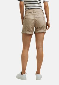 Personne portant un short beige retroussé, des baskets blanches et un t-shirt à manches courtes rayé noir et blanc, debout de dos sur un fond uni.