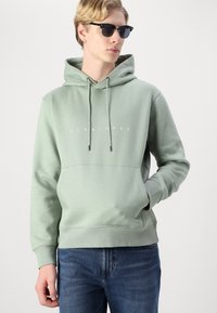 Pullover verde chiaro realizzato in tessuto morbido, con tasca frontale e testo del logo bianco. Abbinato a jeans blu e occhiali da sole neri.