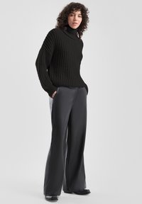 Gebreide zwarte coltrui met een gestructureerd patroon, gecombineerd met zachte, loszittende zwarte broek en platte schoenen.
