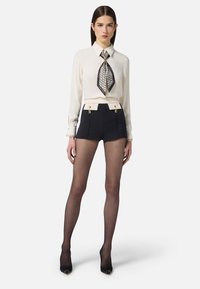 Blusa bianca con sciarpa al collo, pantaloni corti a vita alta blu navy con dettagli dorati, collant neri trasparenti e tacchi alti neri.