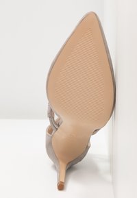 Chaussure à talon haut en suède beige unique avec bout pointu et semelle texturée, inclinée contre un mur et un sol blancs.