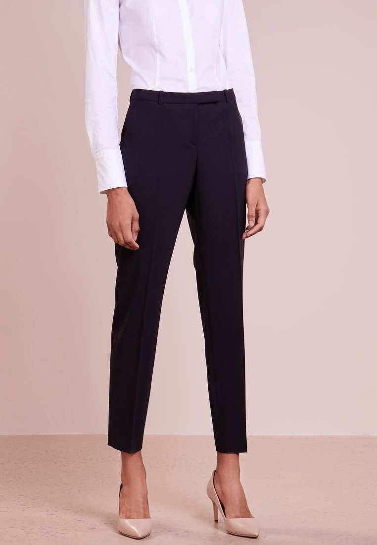 Marineblauwe, op maat gemaakte broek met een slim fit, geplooide design en zijzakken, gecombineerd met een witte blouse en beige hakken.