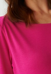 Istuutettu T-paita kirkkaan vaaleanpunaisesta kankaasta, jossa on sileä pinta. Kaula-aukko on pyöreä, ja siinä on lyhyet hihat, joissa on hillittyjä ompelu koristeita.