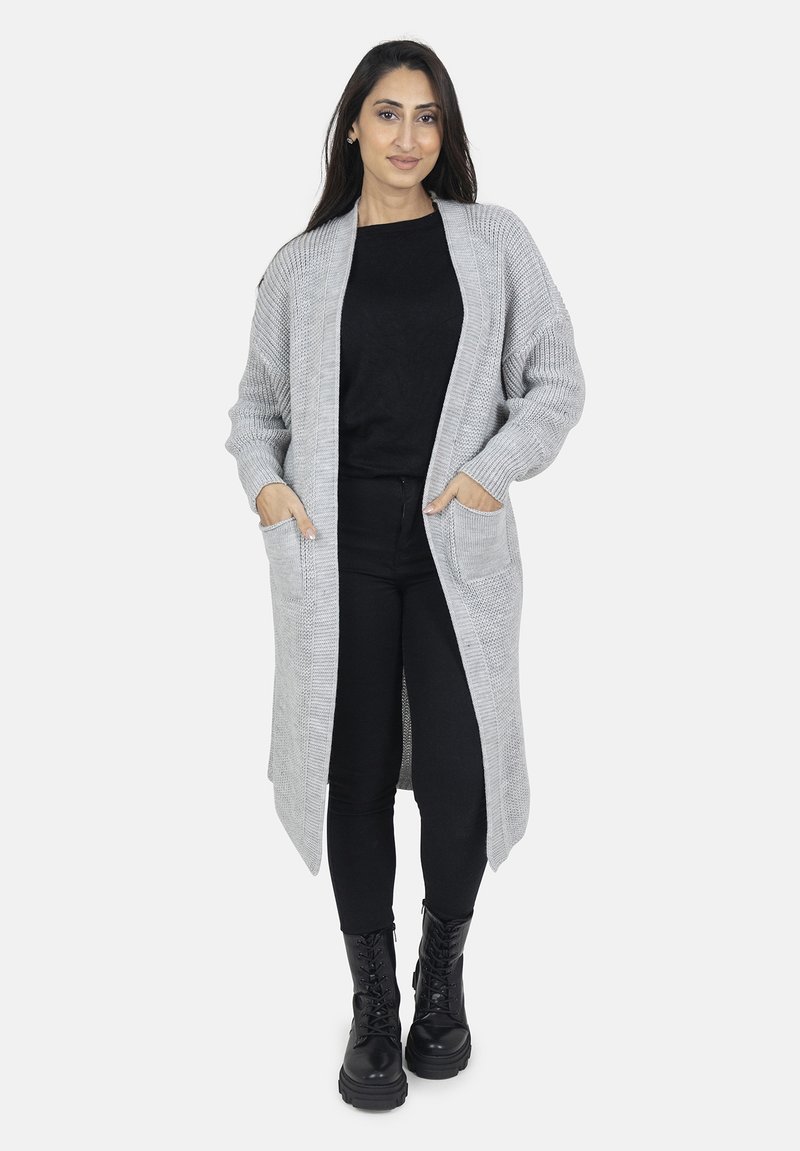 Lange grijze gebreide cardi met een gestructureerd patroon, open voorkant, twee voorzakken en geribbelde manchetten, gecombineerd met een zwarte top en legging.