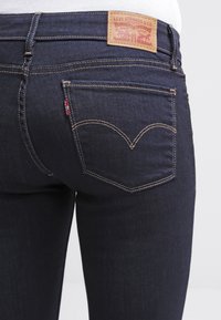Jeans de mezclilla azul oscuro con una cintura alta, que cuentan con bolsillos traseros con un patrón de costura curva distintivo y una etiqueta de cuero con marca.