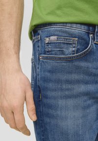 Tamno plave traperice s uskim krojem, koje imaju pet džepova. Tkanina ima blagu teksturu, a vidljiva je mala oznaka s natpisom "s.Oliver".