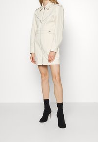 Krátké krémové šaty na zip s dvěma náprsními kapsami a přiléhavým pasem. Dlouhé rukávy s manžetami na knoflíky. Nosič má na sobě černé kotníkové boty.