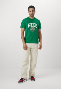 Grøn Nike T-shirt med hvidt logo og cirkulært grafisk design, beige bukser, sorte og røde sneakers, står mod en ensfarvet baggrund.