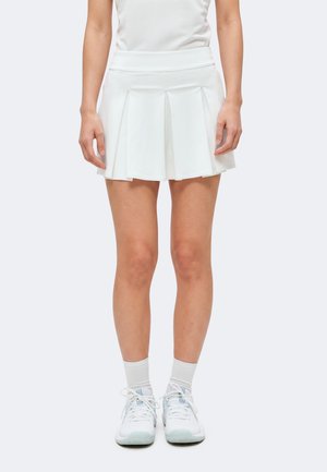 Mujer con falda de tenis blanca plisada, calcetines blancos y zapatillas deportivas blancas, de pie frente a un fondo claro y sencillo.