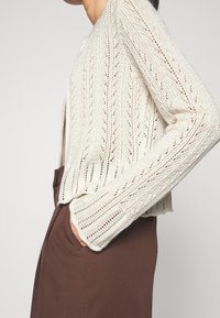 Sweter kardigan w kolorze kremowym, z teksturowanym wzorem jodełki, otwartym przodem i długimi rękawami, zestawiony z brązową odzieżą.