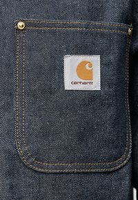 Σκούρο τζιν ύφασμα με διαγώνιο ύφανση, που διαθέτει μια μοναδική τσέπη στο στήθος με λευκή ετικέτα και πορτοκαλί λογότυπο, περιγραμμένη με κίτρινη ραφή.