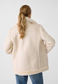 Frau mit langem blonden Pferdeschwanz, die eine beigefarbene Lammfelljacke und blaue Jeans trägt, mit dem Rücken gegen einen schlichten weißen Hintergrund gewandt.