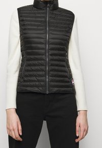 Svart pufferväst med ståkrage, quiltat mönster och dragkedjefickor på sidorna. Bärs över en vit långärmad tröja. Accenterad med logotyp.