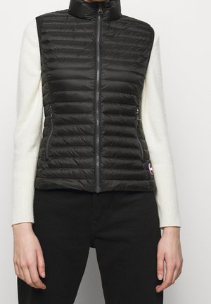 Sort puffervest med ståkrave, quiltet design og lynlåslommer i siderne. Båret over en hvid langærmet sweater. Accentueret med logo.