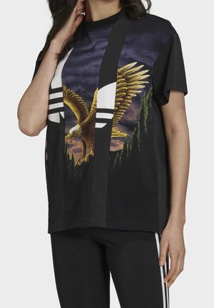 Osoba nosząca czarną koszulkę z dużą grafiką orła i białym logo, w połączeniu z czarnymi spodniami z białymi paskami po bokach.