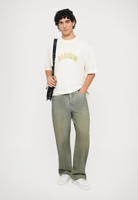 Weißes Baumwoll-T-Shirt in Übergröße mit goldener "BARROW"-Aufschrift, kombiniert mit lockeren grünen Jeans und weißen Sneakers. Schwarze Schultertasche.