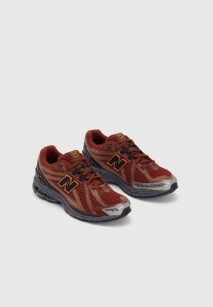 Paar weinrote und schwarze Laufschuhe mit Mesh-Gewebe, schwarzen Sohlen, Schnürsenkeln und orangefarbenem „N“-Logo an den Seiten auf hellgrauem Hintergrund.
