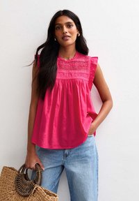 Roze mouwloos topje met ruches op de schouders en kantdetails bij de halslijn, gecombineerd met lichtblauwe spijkerbroek en een gevlochten tas.