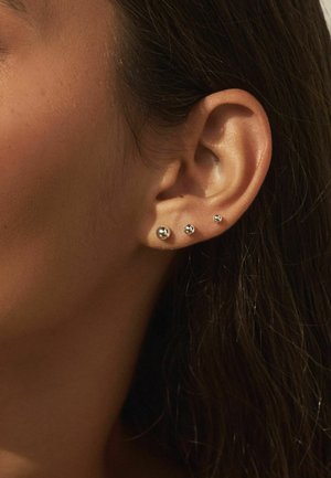 Nahaufnahme eines rechten Ohrs mit vier kleinen silbernen Steckerohrringen, die in abnehmender Größe vom Ohrläppchen nach oben angeordnet sind.