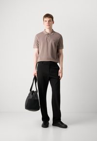 Brun polo skjorte med kort erme, svarte bukser og svarte sneakers. Holder en grå veske, står på en ensfarget bakgrunn.