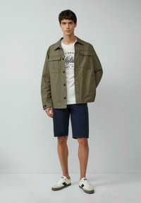 Oliwkowa, zapinana na guziki kurtka z dwoma kieszeniami na piersi, biała koszulka z nadrukiem, granatowe szorty oraz białe trampki z brązowymi akcentami.