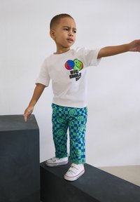 Fehér póló színes mosolygós arcokkal és a "NAGY MOSOLY NAP" felirattal, kék kockás nadrággal, amelyen mosolygó minták láthatók, fehér sportcipővel.