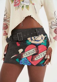 Minifalda de faux leather negra con gráficos coloridos de flores y corazones, con el texto "AMOR ETERNO" y un cinturón negro desmontable.