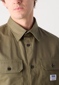 Olive green button-up shirt with a pointed collar, two chest pockets, black buttons, and a small blue and white label on the lower side.
