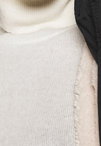Pull à col roulé en maille côtelée crème associé à une veste noire, avec une doublure en shearling texturée le long du col et des bords.