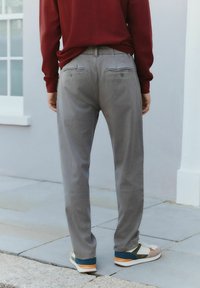 Graue Hose mit geradem Schnitt, zwei Gesäßtaschen mit Knopfverschluss, getragen mit einem kastanienbraunen Oberteil und mehrfarbigen Sportschuhen.