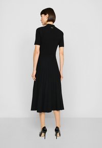 Svart plisserad klänning med korta ärmar, ribbad textur, figursydd överdel, utsvängd kjol och en liten loggdetalj på baksidan. Svarta klackskor.